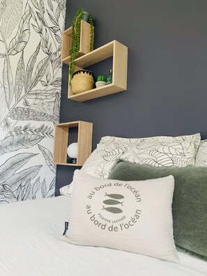 A bedroom with a dark wall, a leaf-patterned wallpaper, a bed with a green pillow, and two wooden shelves with decorative items.