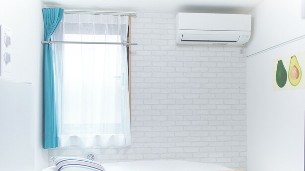A small, bright room with a bed, a window with curtains, an air conditioner, and a wall with a picture of avocados. - Zenith Shinjuku Hinoki Building B (Tokyo)