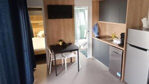 A compact hotel room with a bed, a small dining table with chairs, a kitchenette with a refrigerator, and a television mounted on the wall.