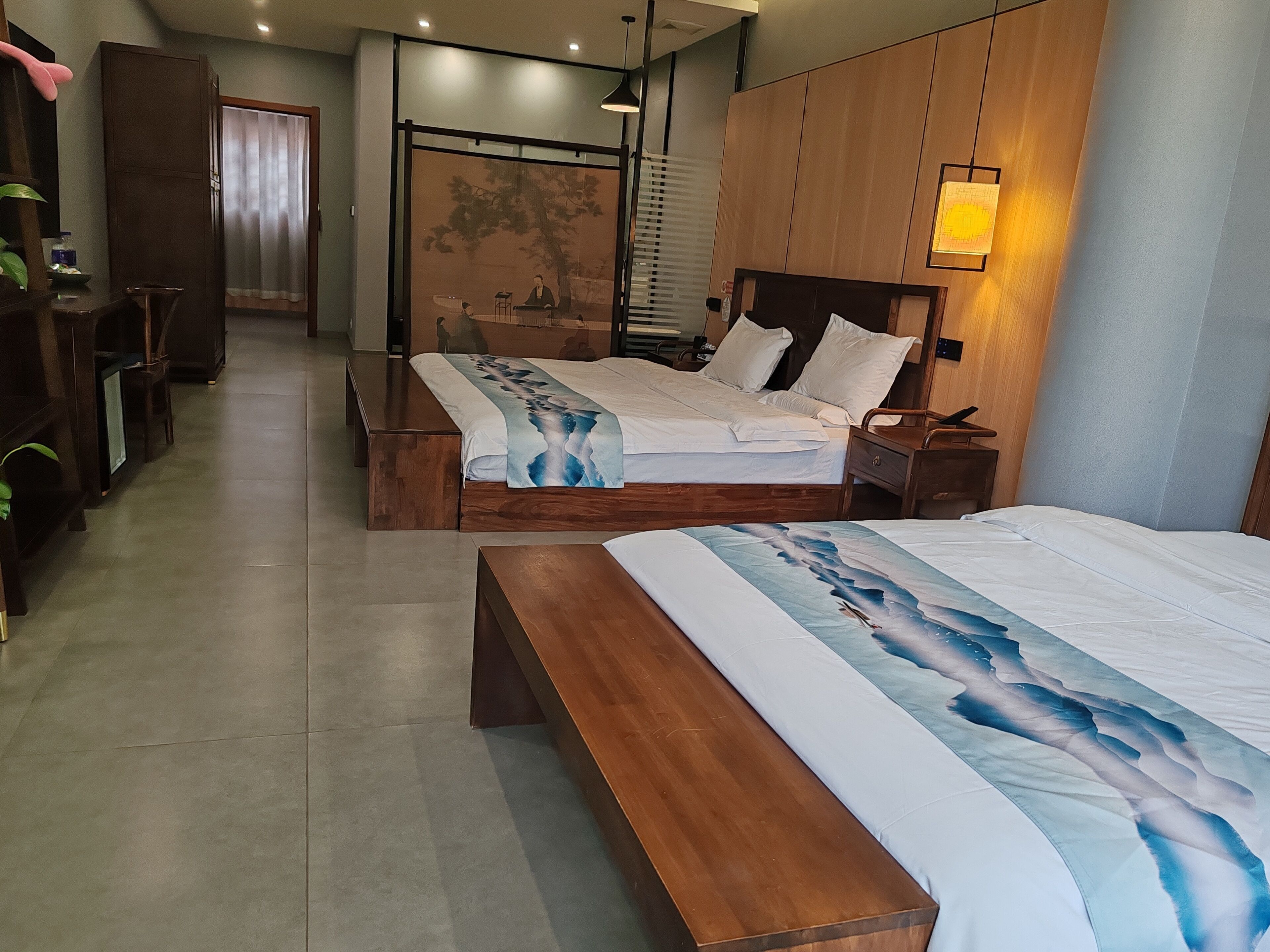 A modern hotel room with a large bed, a smaller bed, a wooden bench, a desk, and a chair.