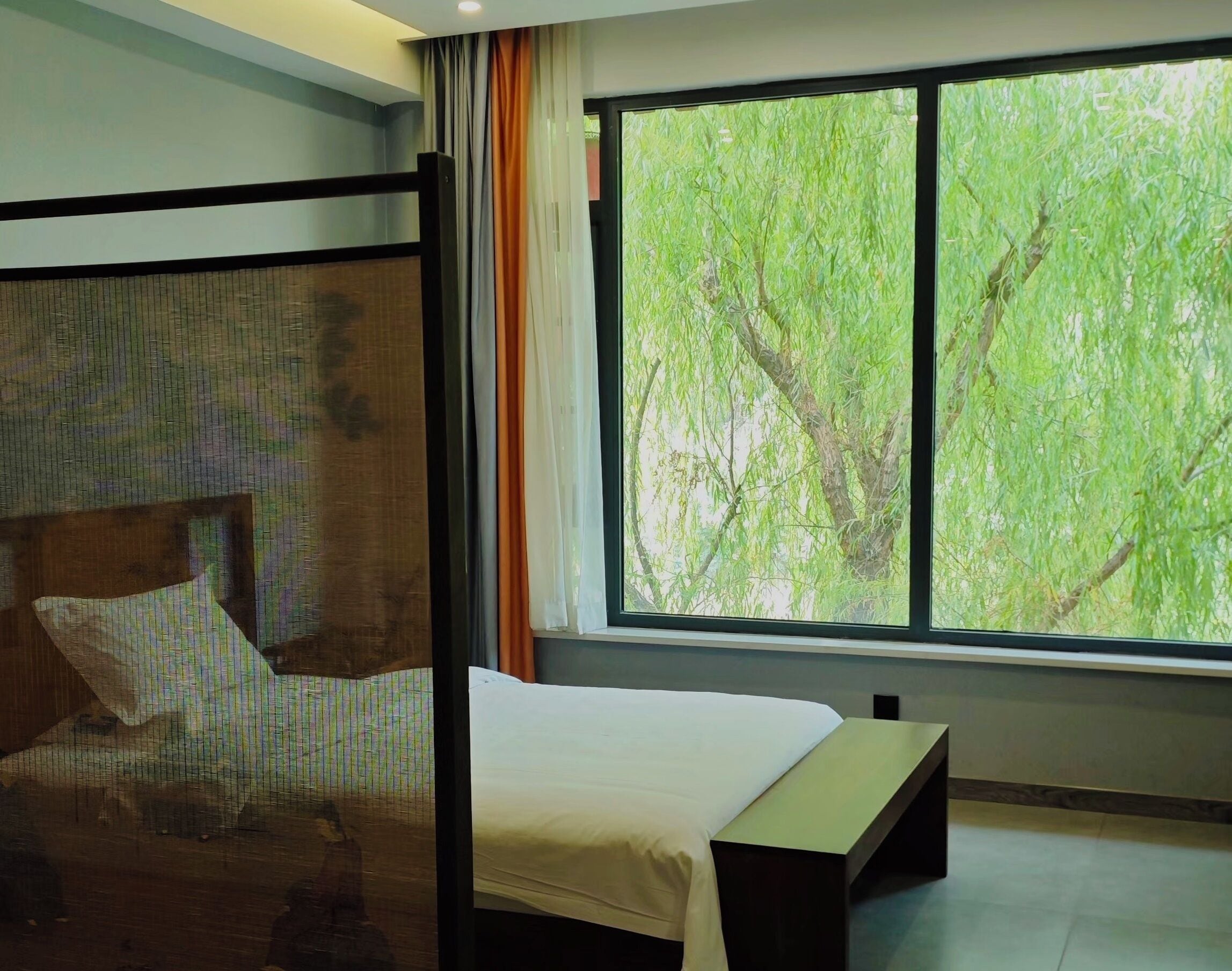 A bedroom with a large window, a bed, a bench, and a wall with a textured design.