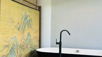 A bathroom with a black freestanding bathtub, a white sink, and a textured wall with a landscape painting.