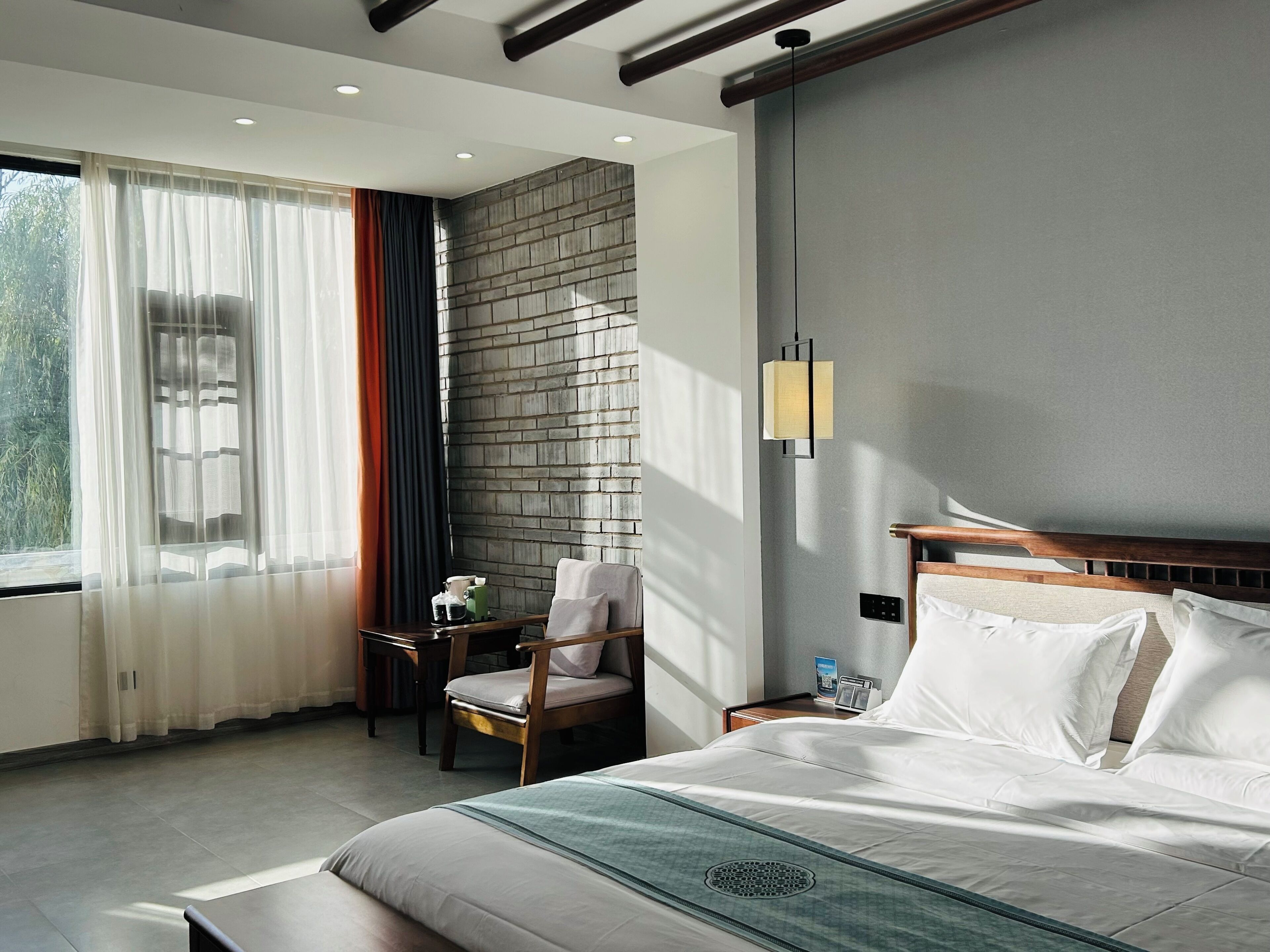A hotel room with a bed, a chair, a small table, a wall-mounted lamp, and a window with curtains.