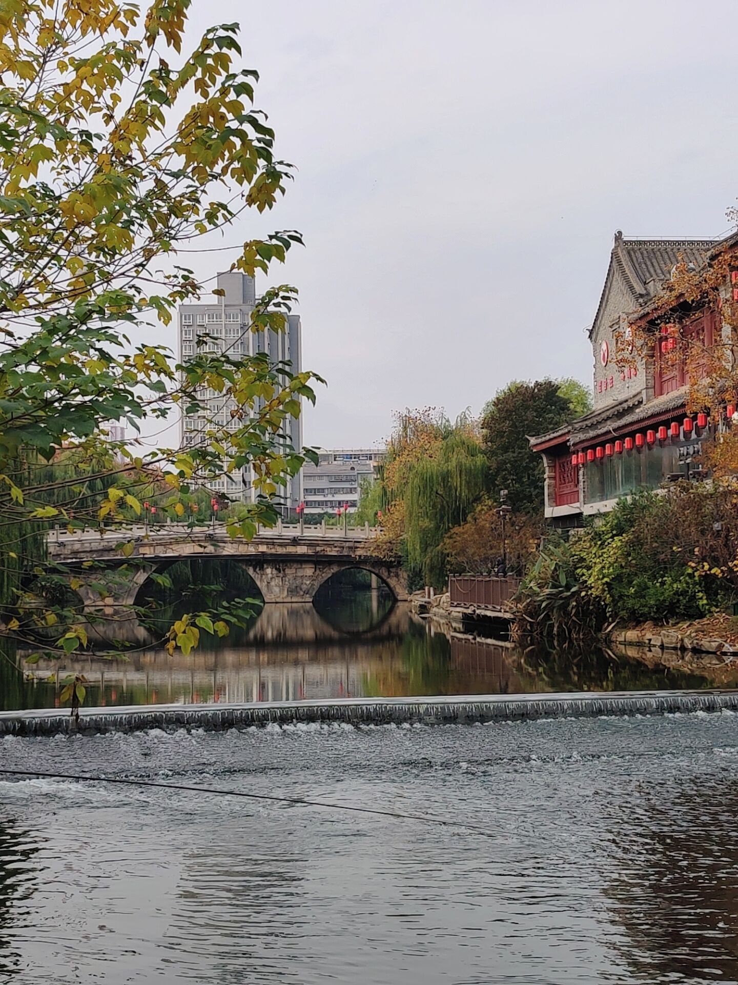 A traditional building with a tiled roof and red lanterns, a stone bridge over a calm river, and modern high-rise buildings in the background.