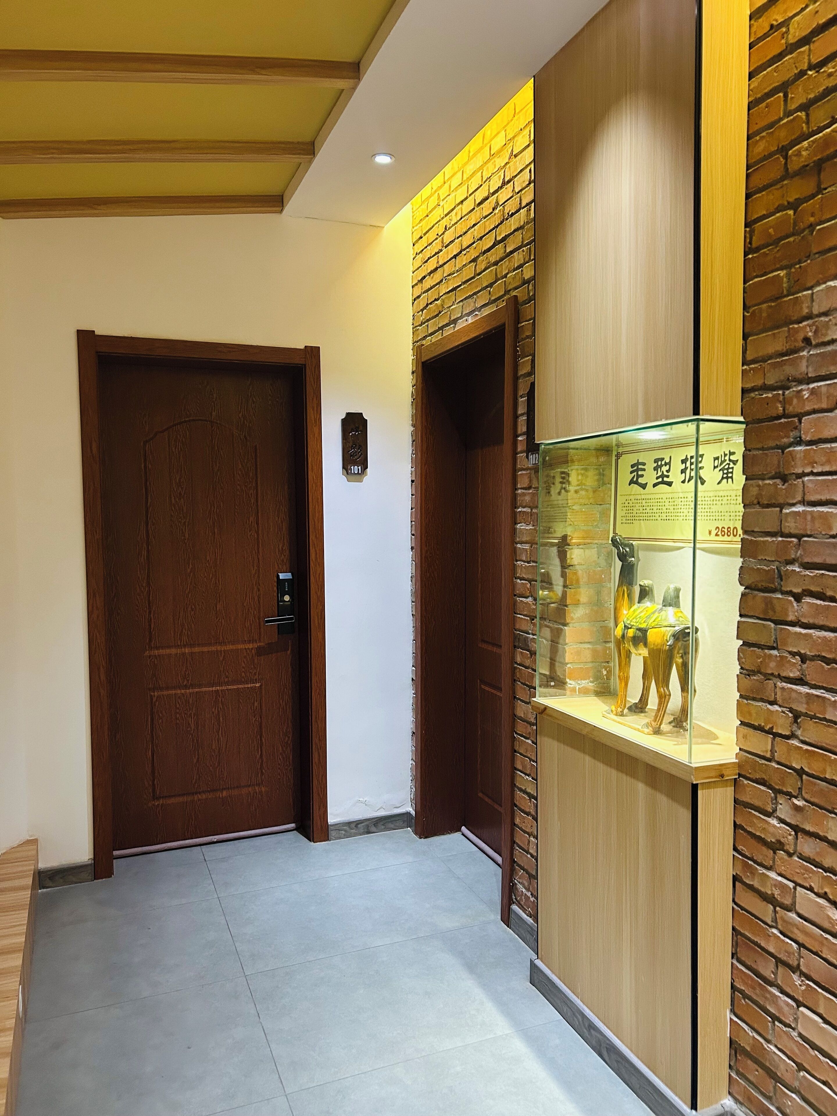 Un pasillo con una puerta de madera, una placa con número y una vitrina de cristal.