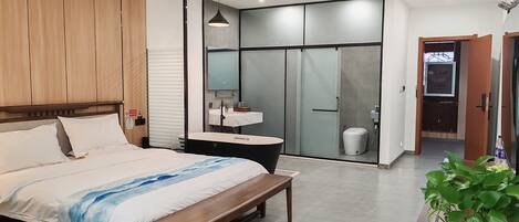 Una habitación de hotel moderna con cabecera de madera, una cama grande con colcha blanca y azul, un banco de madera, una pequeña mesa redonda, un baño con puerta de vidrio y una planta en maceta.