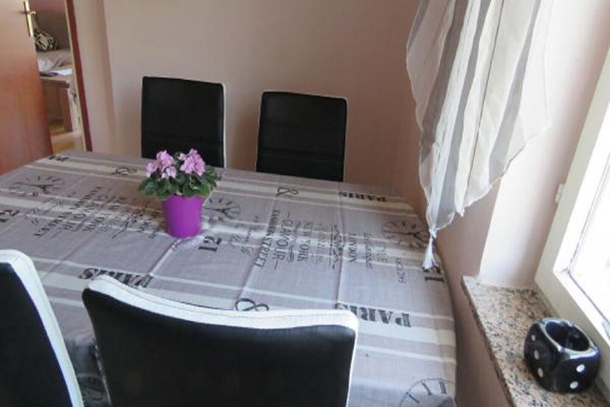 A dining area with a table covered by a newspaper print tablecloth, two black chairs, a purple flower vase, and a window with curtains.