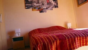 Une chambre d’hôtel avec un lit recouvert d’une couverture rouge, une table de chevet avec une lampe et un tableau représentant un paysage urbain accroché au mur.