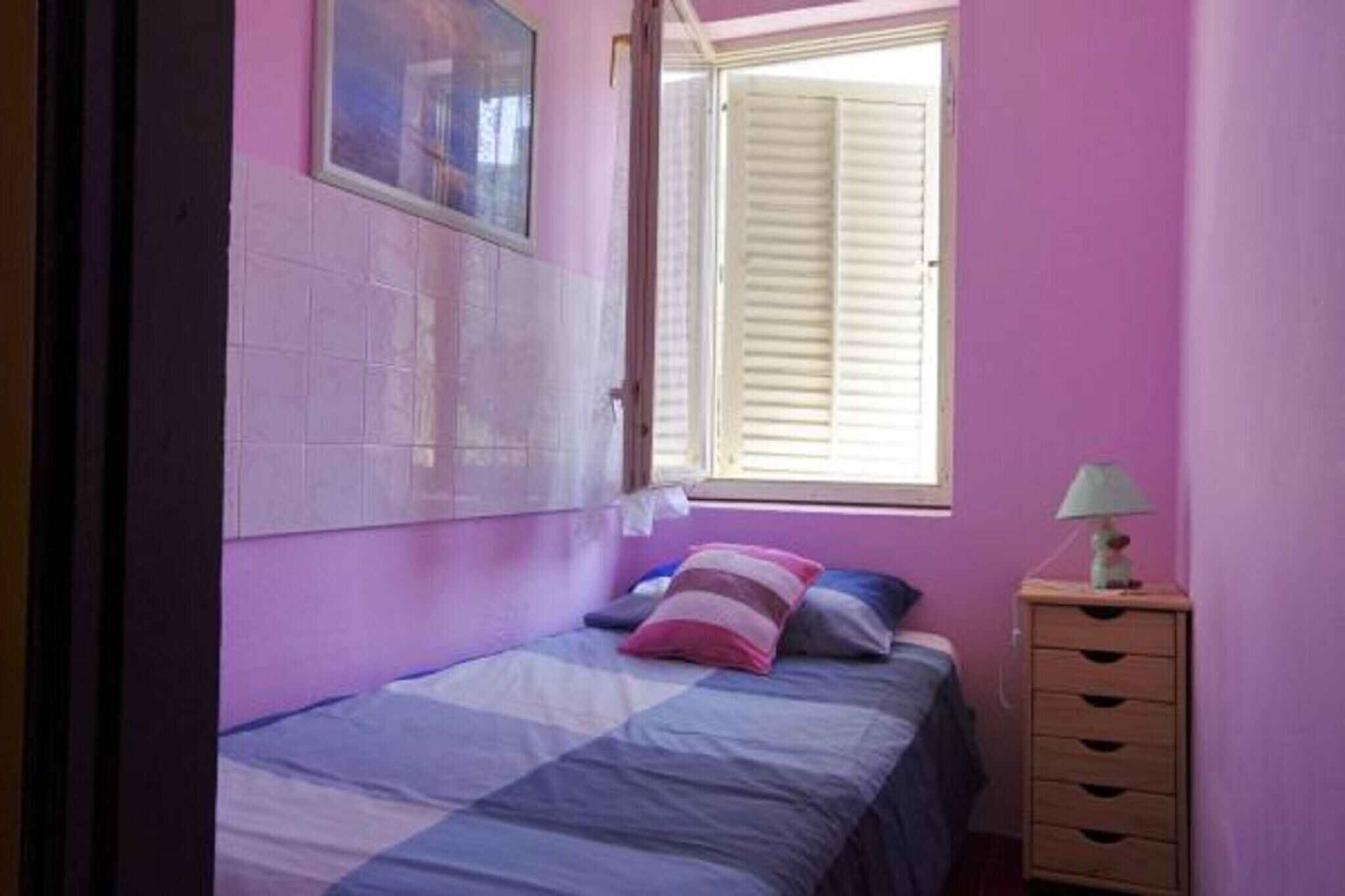 Une chambre à coucher avec un lit, une fenêtre avec des stores, une commode et une lampe.