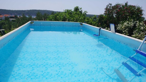 Une piscine rectangulaire dont le fond est recouvert de carrelage, entourée de verdure et surplombée d’un ciel dégagé.