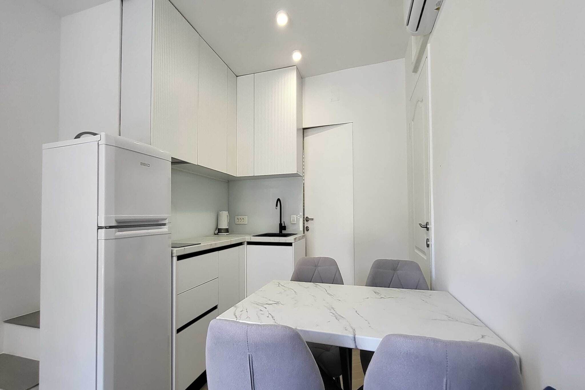 Een kleine keuken met witte kasten, een inbouwkoelkast, een spoelbak en een marmeren eettafel met vier stoelen.