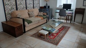 A living room with a sofa, coffee table, and a small wooden chest. There is a desk with a computer and a chair. A decorative screen is behind the sofa. - T3 apartment (two bedrooms) on garden level, garden 120 m2 town center, (Saint-Jean-de-Luz)