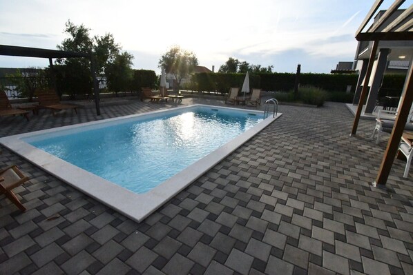 A rectangular swimming pool with a tiled deck, surrounded by lounge chairs and umbrellas.