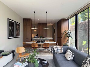A modern living room with a sofa, coffee table, and chairs, a dining area with a round table, and a kitchen with wooden cabinets and pendant lighting.
