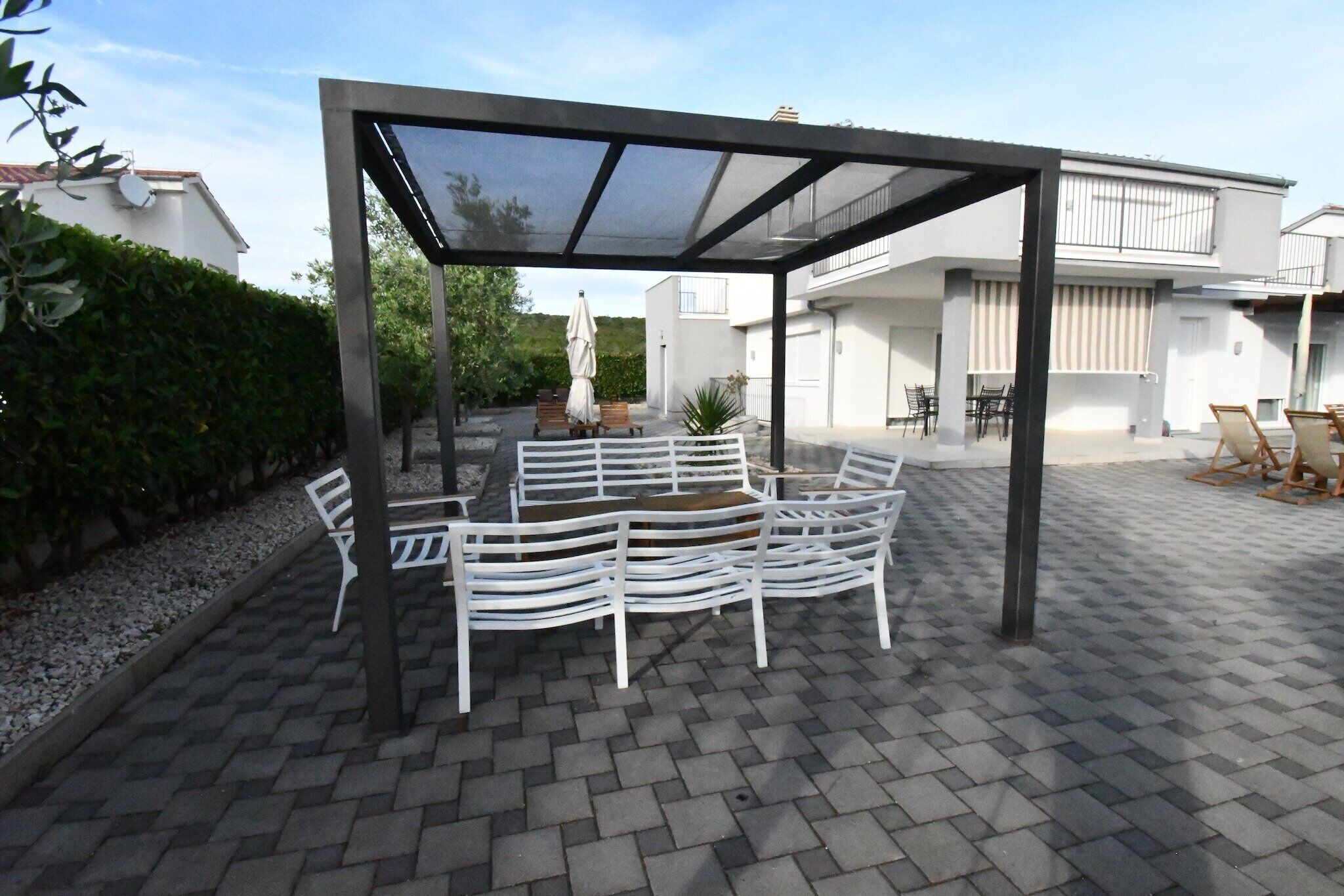 Ein überdachter Außenbereich mit weißen Metallmöbeln, einer Pergola und einer gepflasterten Terrasse.