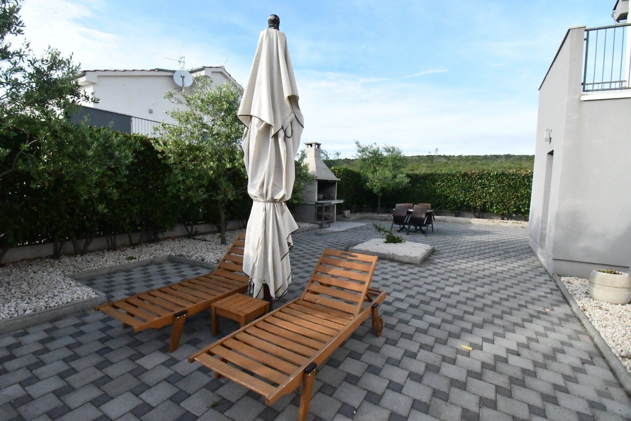 Un patio con dos sillones de madera y una sombrilla, rodeado de un seto y un edificio con antena parabólica.