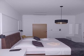 A hotel room with a bed, a bedside table, a remote control, a door, and a hanging light fixture.
