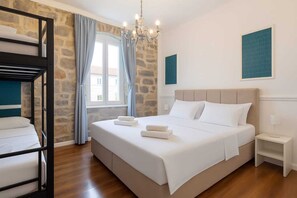 A bedroom with a bunk bed, a large bed, a chandelier, a window with curtains, and a nightstand with a lamp.