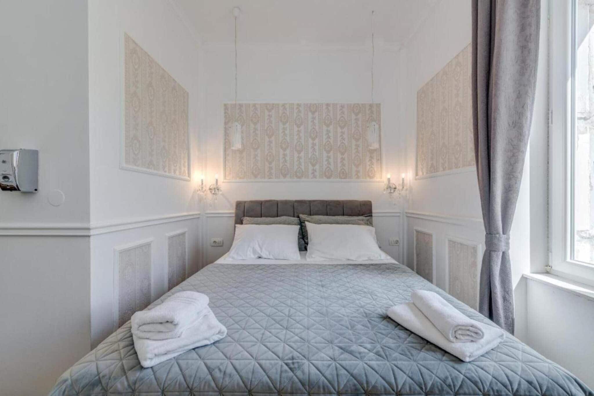 Habitación de hotel con cama, dos apliques en la pared, una ventana con cortinas y un termostato en la pared.