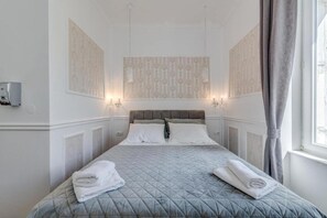 A hotel room with a bed, two wall-mounted sconces, a window with curtains, and a wall-mounted thermostat.