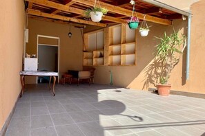 Un espacio cubierto con piso de baldosas, una mesa de madera, sillas, macetas colgantes y una planta en maceta.