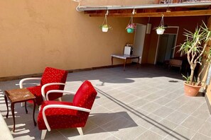 Un patio con sillas rojas, una mesita pequeña, una planta en maceta y jardineras colgantes.