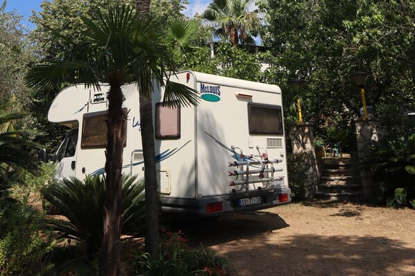 A white RV with 'McLUS' branding parked in a wooded area, surrounded by trees and a stone staircase.