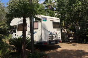 A white RV with 'McLUS' branding parked in a wooded area, surrounded by trees and a stone staircase.
