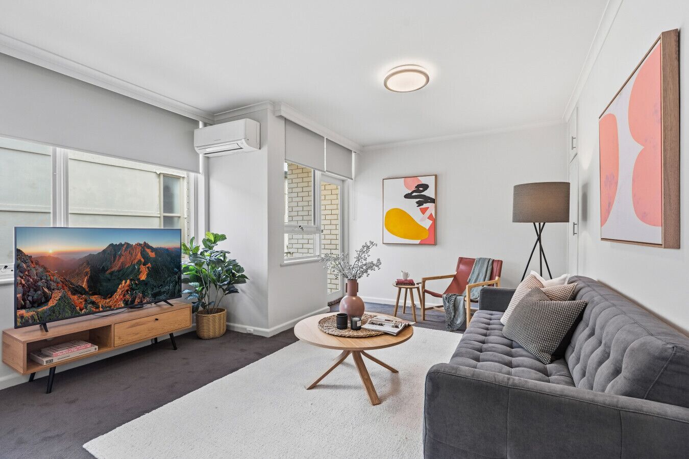 A modern living room with a flat-screen TV, a grey sofa, a coffee table, and a large window.