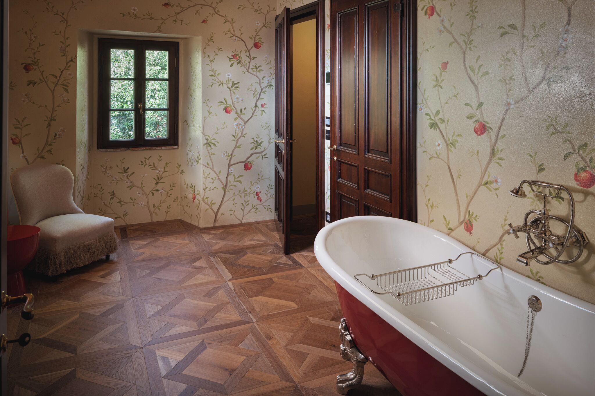 Un baño con suelo de madera, una bañera roja, una puerta de madera y una ventana con vistas a la vegetación.