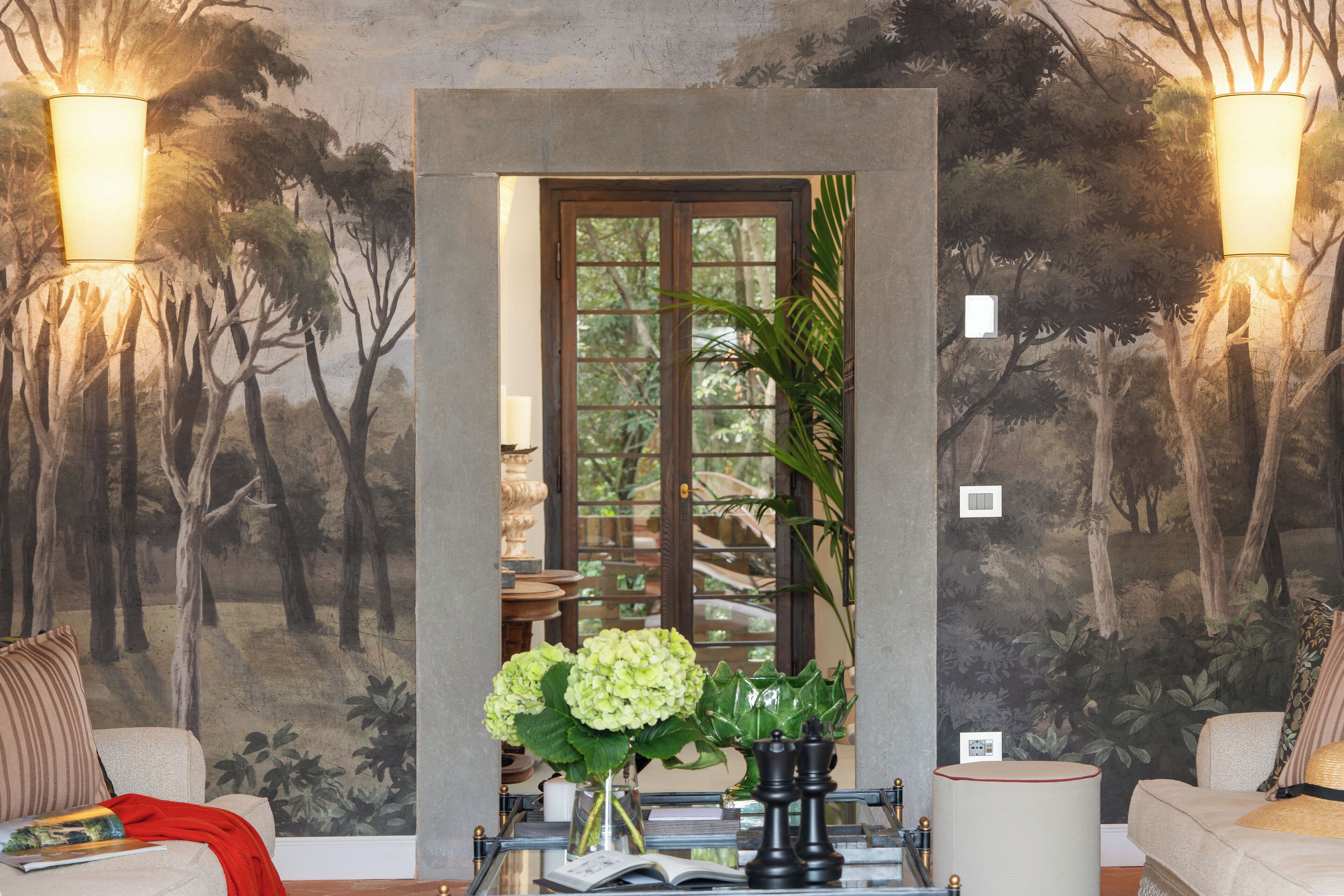 A room with a mural of trees, a wooden door, a vase with flowers, and a lamp.