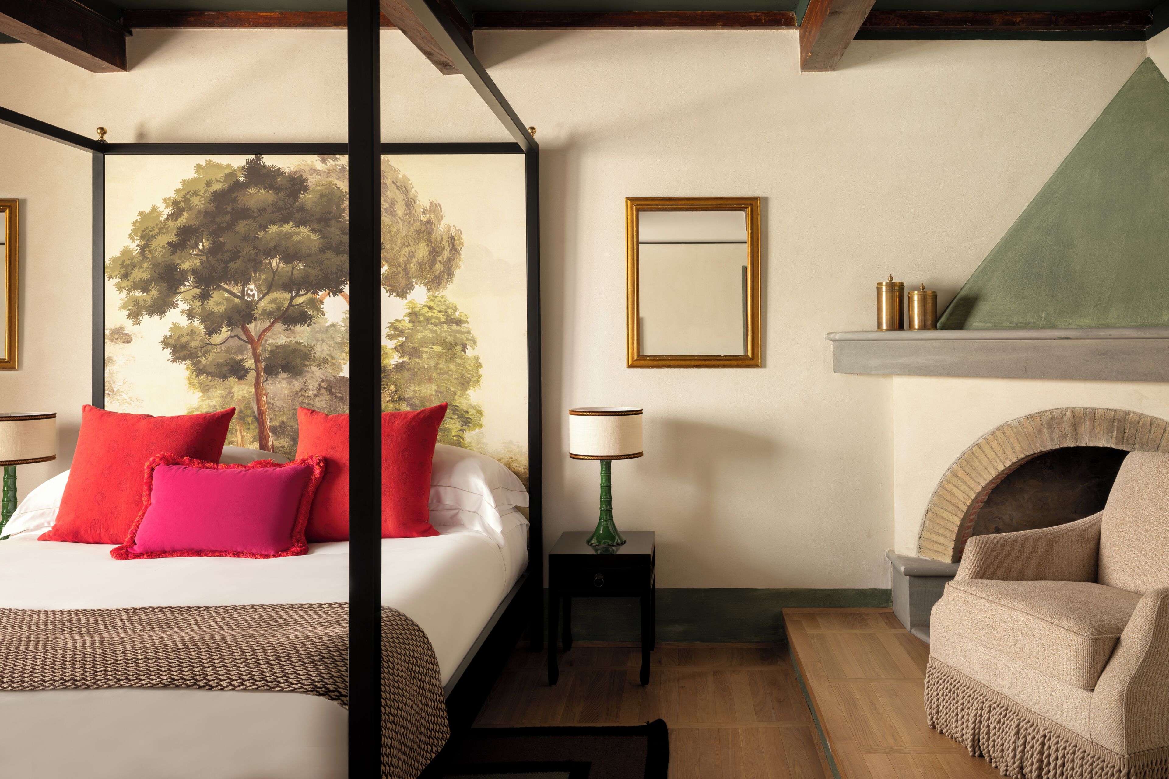 A bedroom with a four-poster bed, a fireplace, a green lamp, and a view of trees through the window.