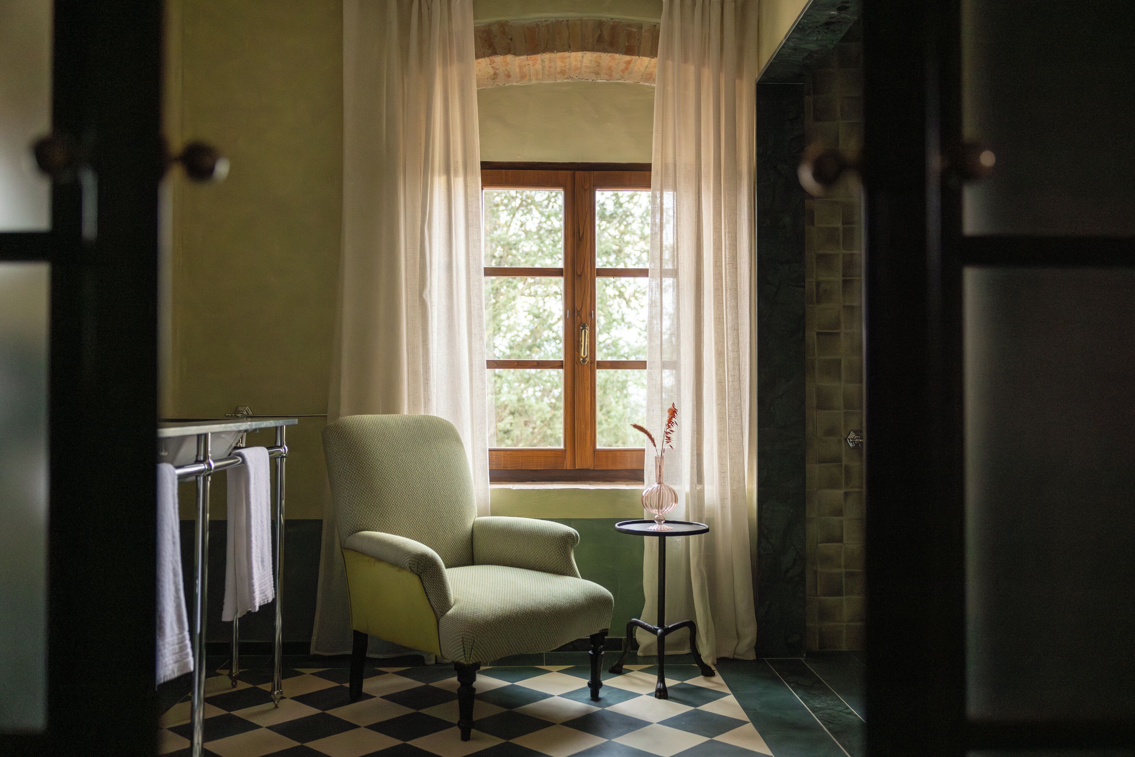 A room with a checkered floor, a green armchair, a small table with a vase, and a window with curtains.