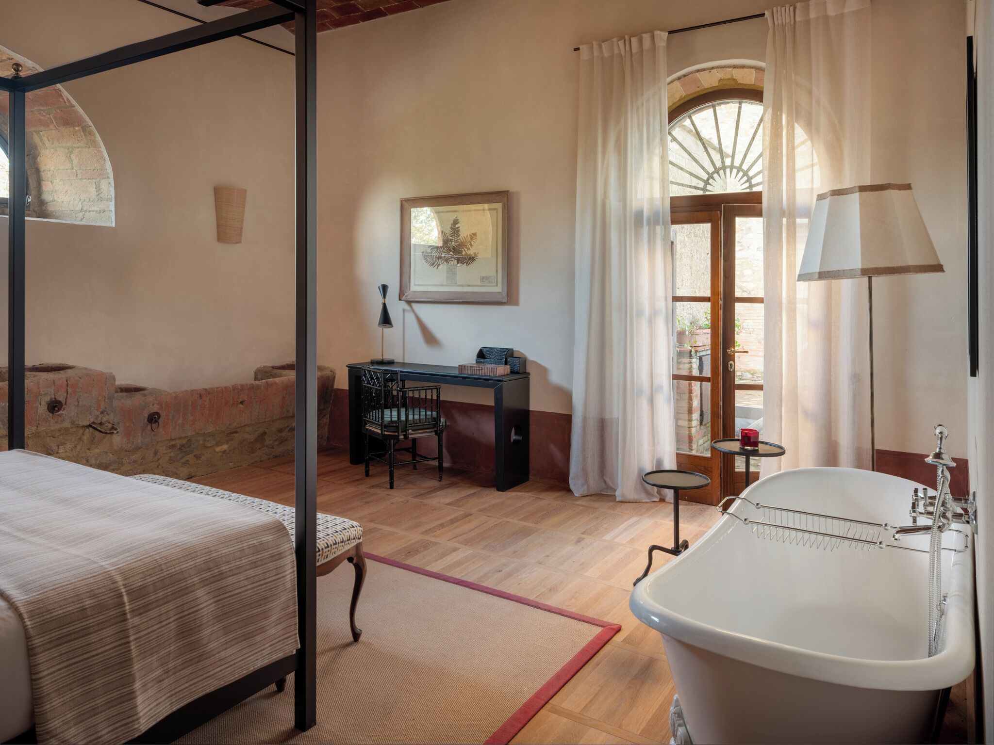 A bedroom with a four-poster bed, a desk with a chair, a large window with curtains, and a white bathtub.
