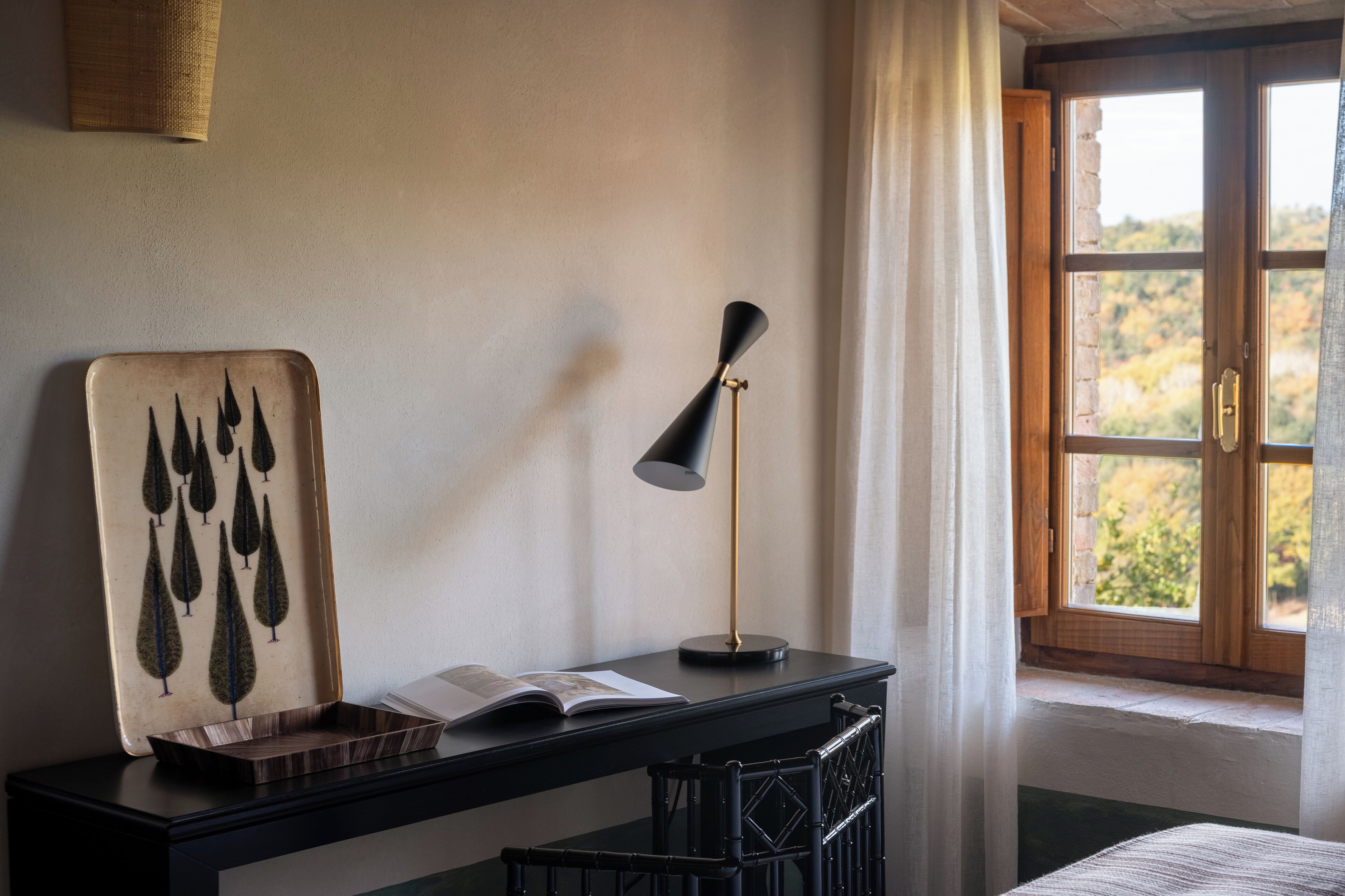 A room with a wooden desk, a lamp, a book, and a window with a view of trees.