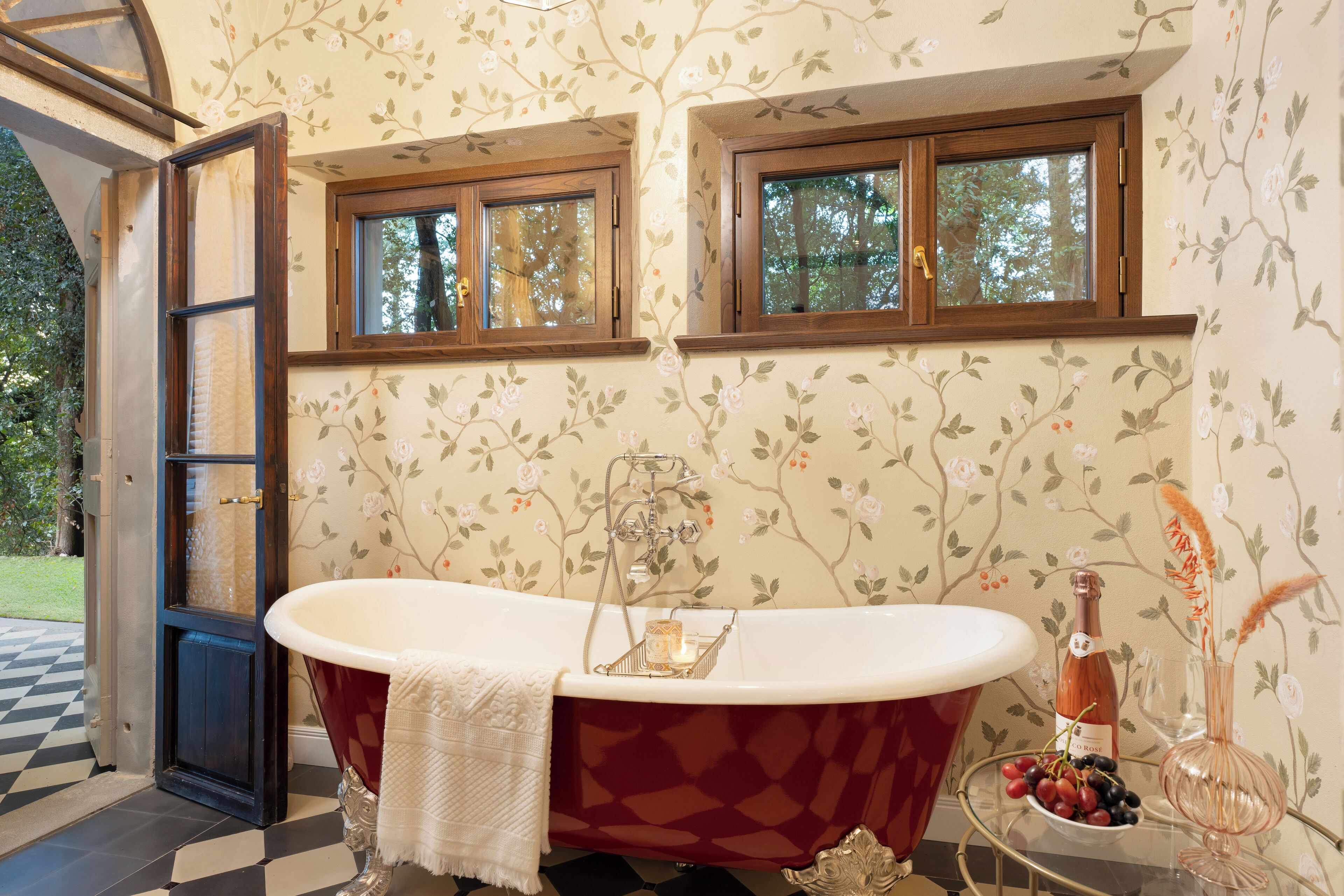 A bathroom with a freestanding tub, two windows, floral wallpaper, and a bottle of wine on a side table.