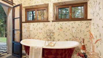 A bathroom with a freestanding tub, two windows, floral wallpaper, and a bottle of wine on a side table.