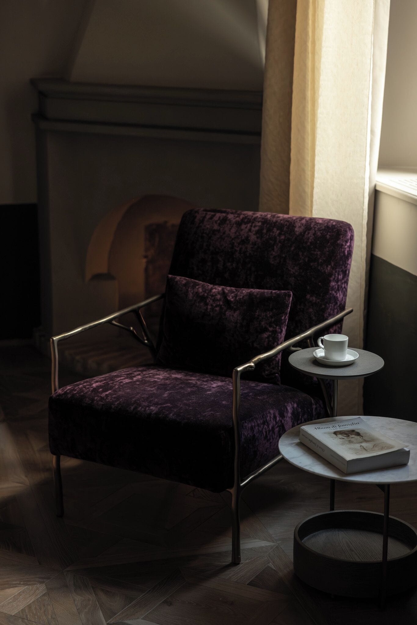 Una habitación oscura con un sillón de terciopelo púrpura, una pequeña mesa redonda con un libro y una taza, y una chimenea al fondo.