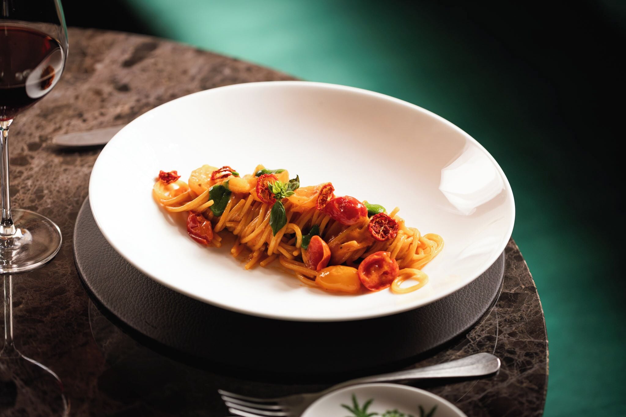 A white plate of pasta with red chili peppers and basil on a dark marble table.