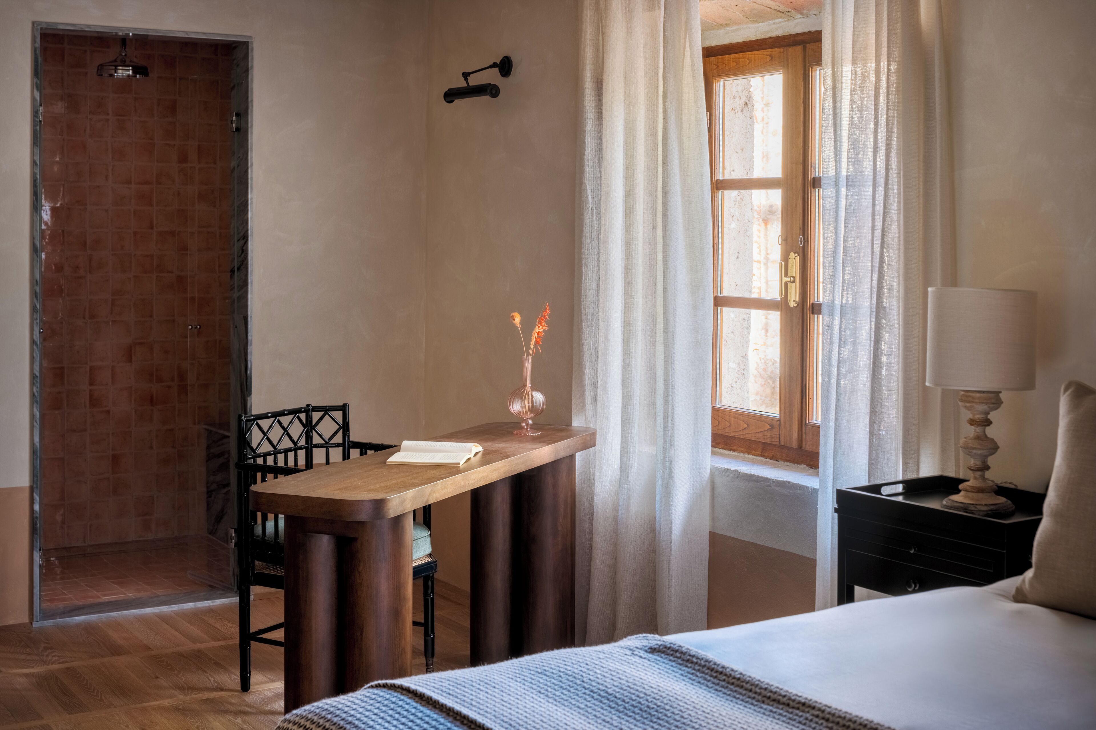 A bedroom with a wooden desk, a chair, a bed, a nightstand, a lamp, and a window with curtains.