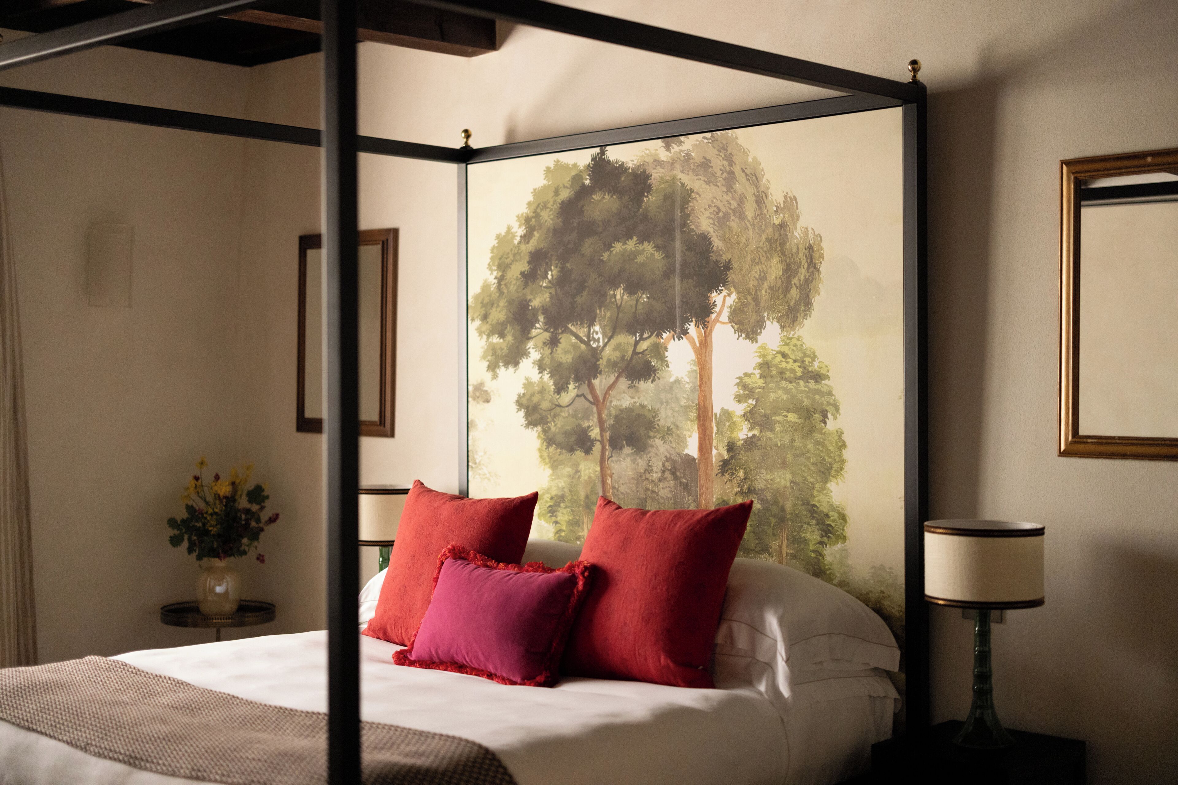 Un lit à baldaquin avec des oreillers rouges, un vase rempli de fleurs et un tableau encadré accroché au mur.