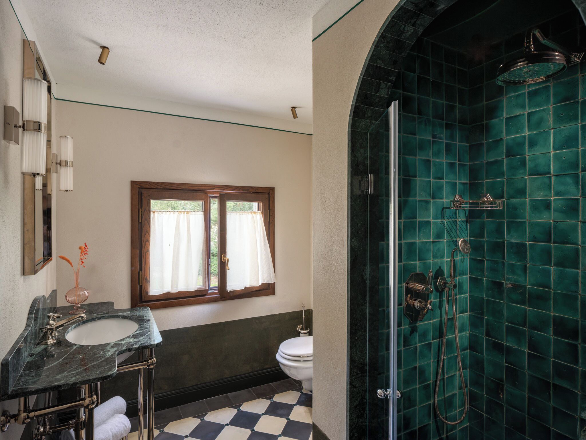 Un baño con lavamanos de mármol, inodoro, ducha con azulejos verdes y ventana con cortinas.