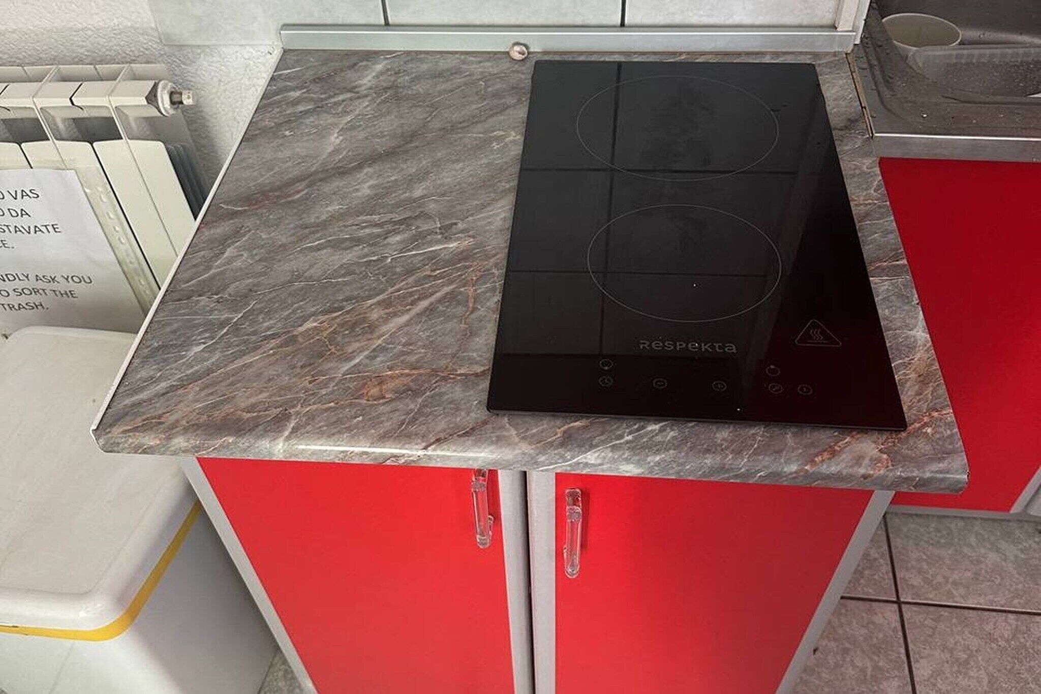 A kitchen with a black induction cooktop on a marble countertop, red cabinets, and a white trash bin.