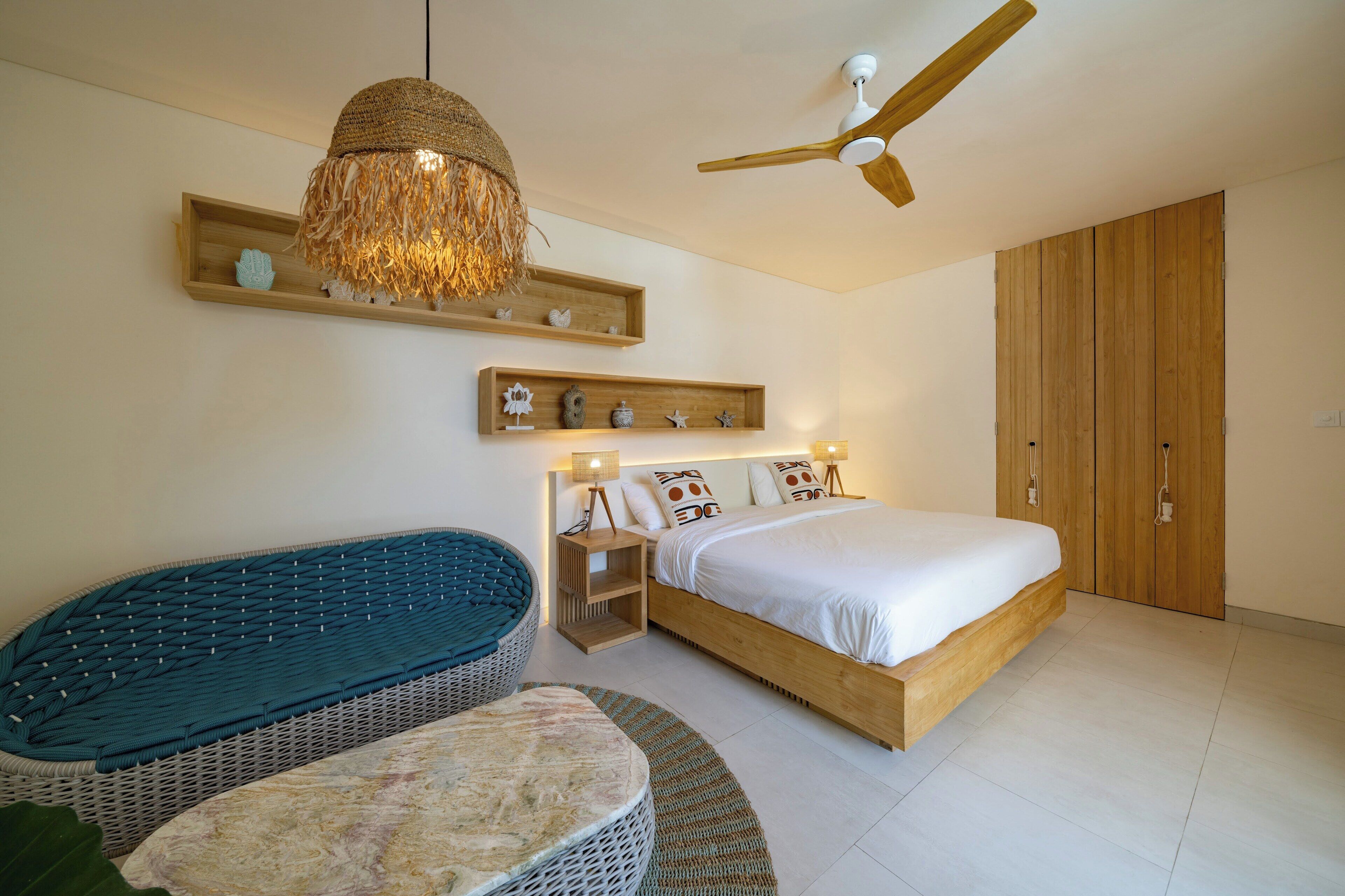 A bedroom with a bed, a ceiling fan, a hanging light fixture, wooden shelves, and a wicker bench.