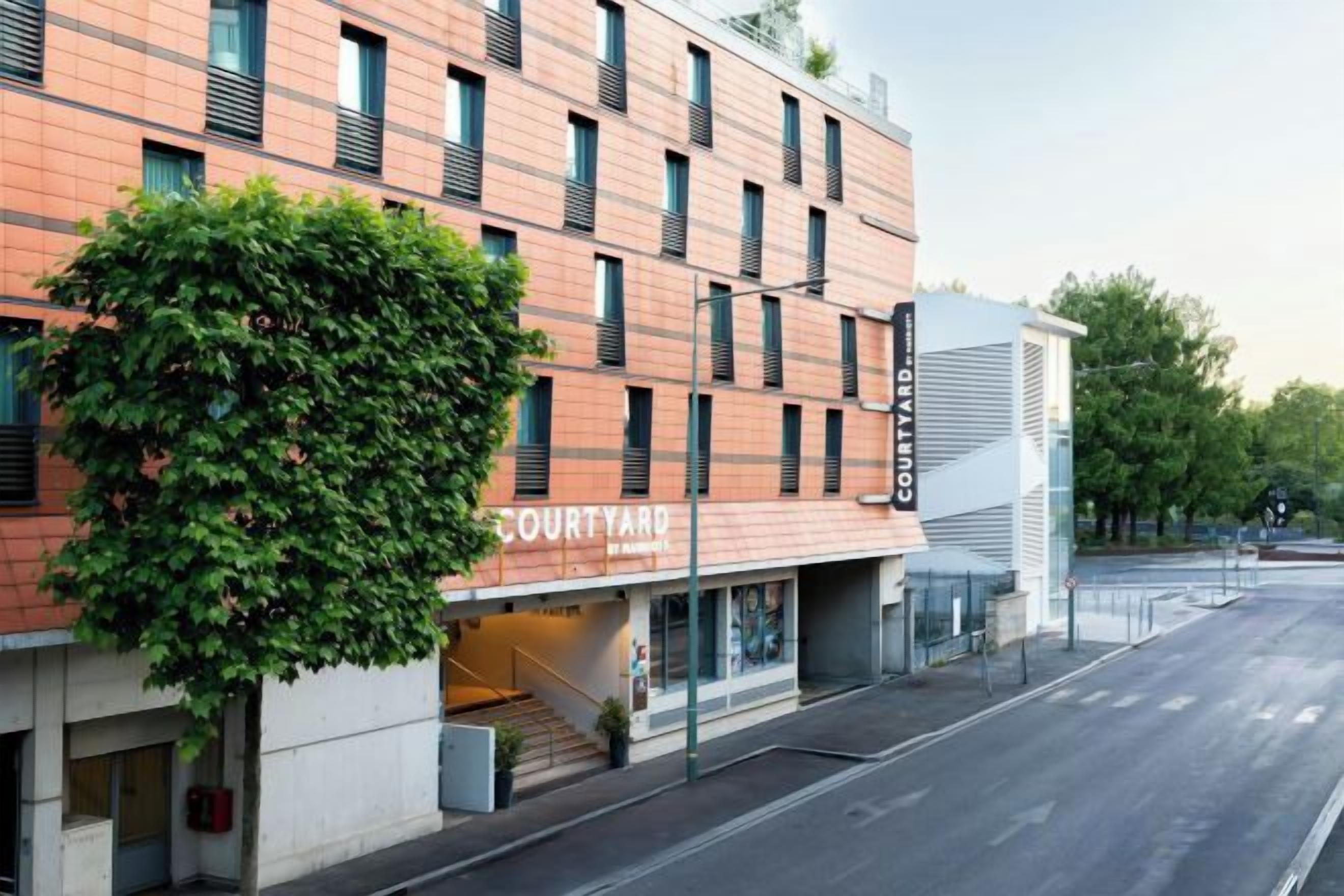 Un immeuble à plusieurs étages, avec une entrée donnant sur une cour intérieure, une façade en briques et un arbre verdoyant à l’avant.