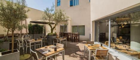 En udendørs terrasse med borde og stole, en pergola og en bygning med store vinduer.