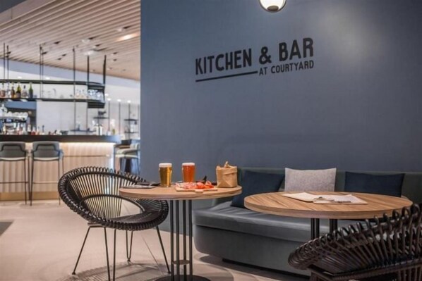 A modern bar area with a sign reading 'KITCHEN & BAR AT COURTYARD,' featuring a bar counter, seating area with a sofa and chairs, and a wall-mounted sign. - Courtyard Paris Arcueil (Arcueil)