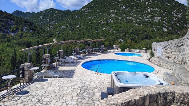 Un área de piscina con dos piscinas, sillas de descanso y un jacuzzi, rodeado de muros de piedra y un paisaje montañoso.