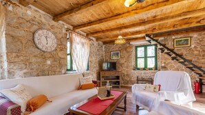 Un salón acogedor con paredes de piedra, vigas de madera, una chimenea de piedra, un televisor y una escalera.