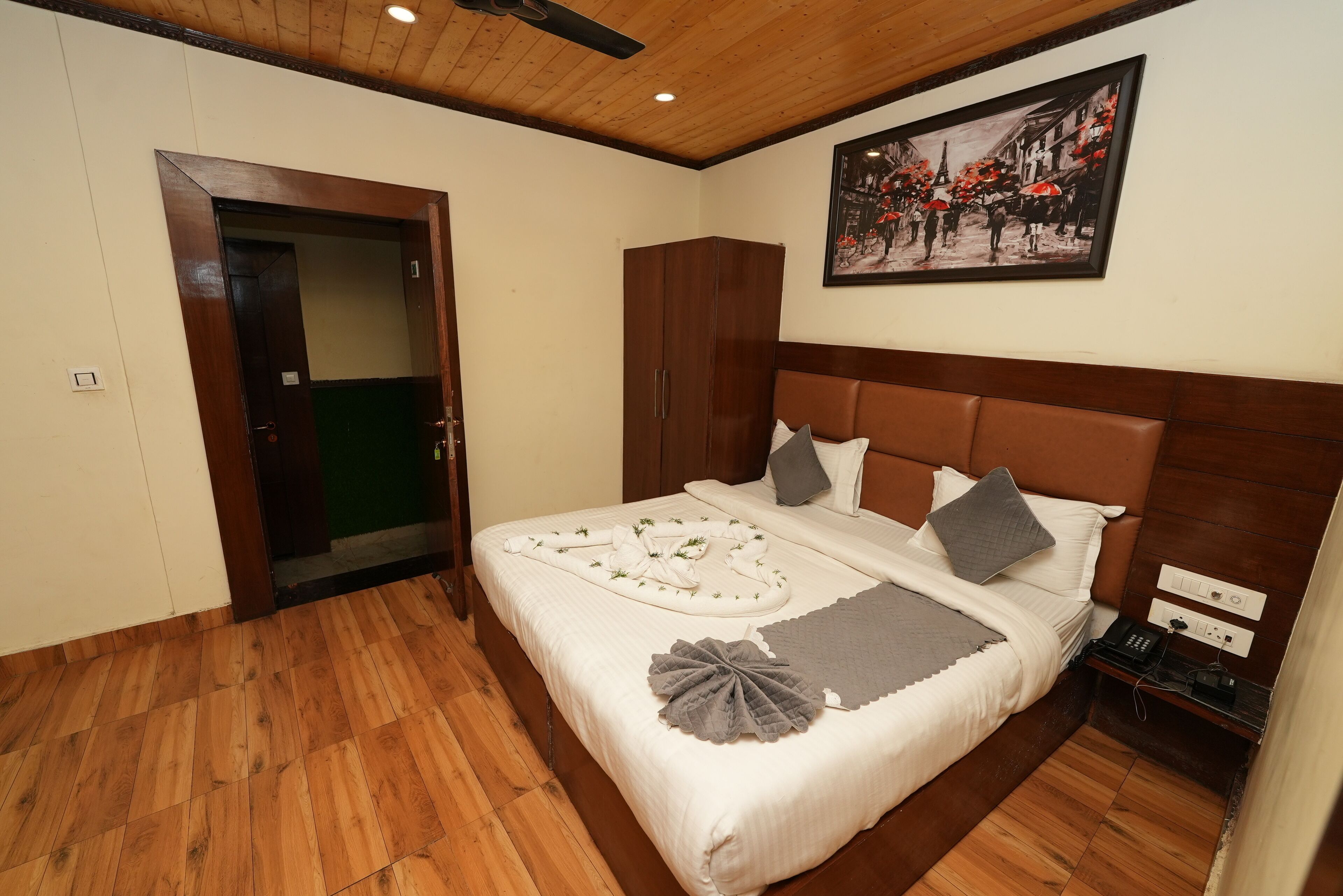 A neatly arranged bedroom with a wooden headboard, a bed with white linens, a grey throw, and a decorative leaf. A wooden wardrobe and a painting are on the wall.